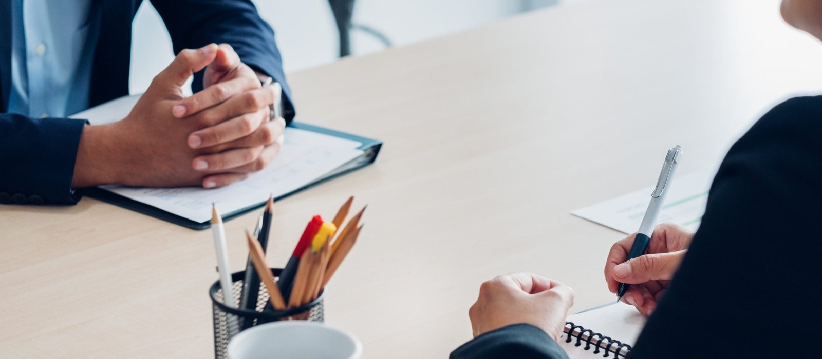 Two people in dark suits sit across a wooden table, one with clasped hands over a clipboard and the other holding a pen above a spiral notebook, beside a pencil holder and a white mug.