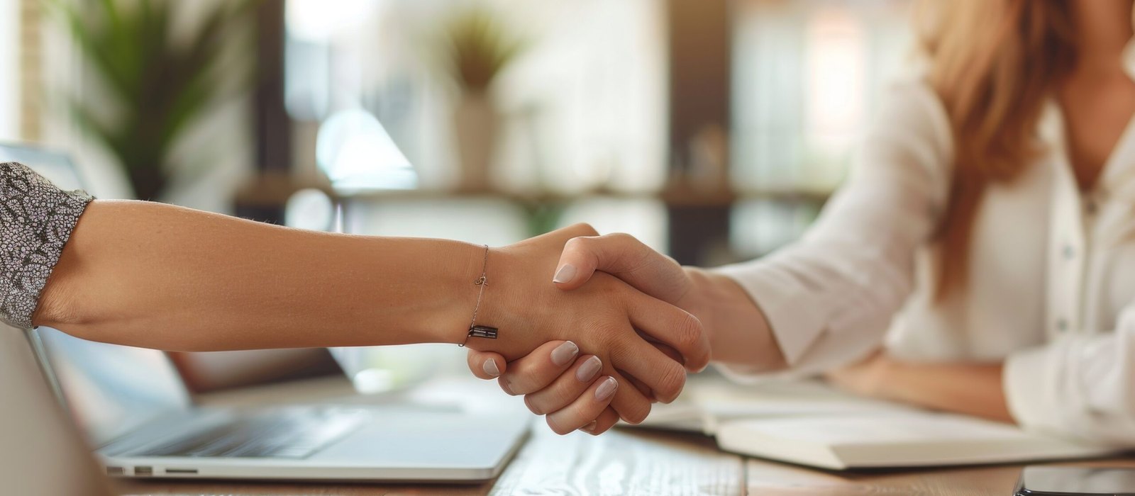 Two people shake hands across a wooden desk with a laptop, notebook and phone nearby in a bright office.