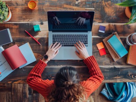 Overhead view of a person working at a laptop on a wooden desk surrounded by notebooks and stationery.
