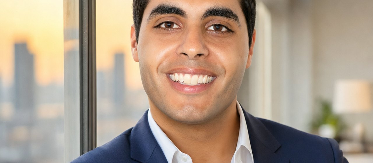 A smiling man in a navy blazer and open-collar white shirt stands beside an office window overlooking a city skyline.