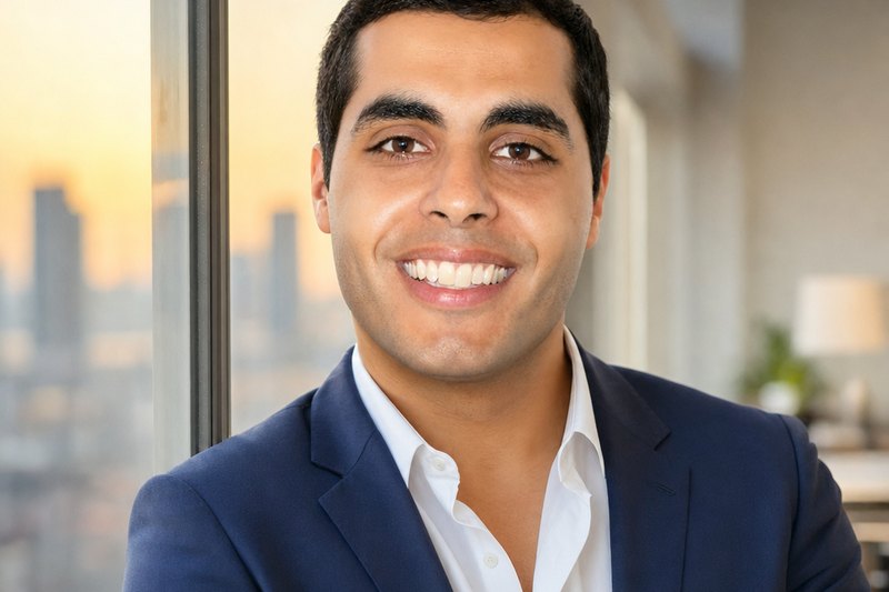 A smiling man in a navy blazer and open-collar white shirt stands beside an office window overlooking a city skyline.